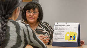 The picture shows an interaction between an American Indian health educator and a teen mother during a parenting lesson. The home-visiting educator uses a tabletop flip chart depicting a typical parenting situation to engage the mother in a discussion about what steps are required to address or resolve the situation.