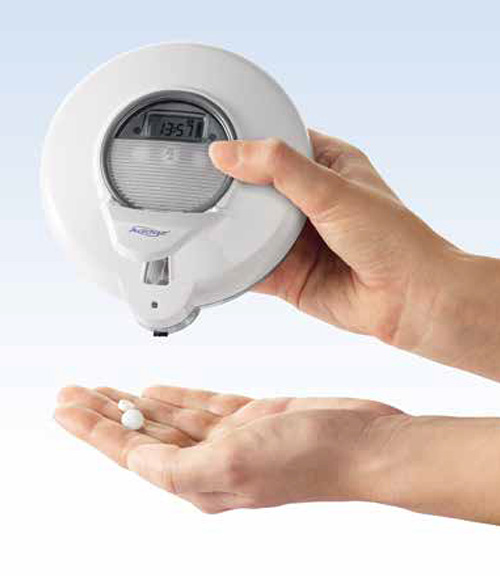The figure shows a hand holding the e-pill Med-O-Wheel device, with a second hand holding two pills underneath. The device is a round box with a dispensing mechanism protruding on one side. In the center, the device has an electronic timer and three buttons.